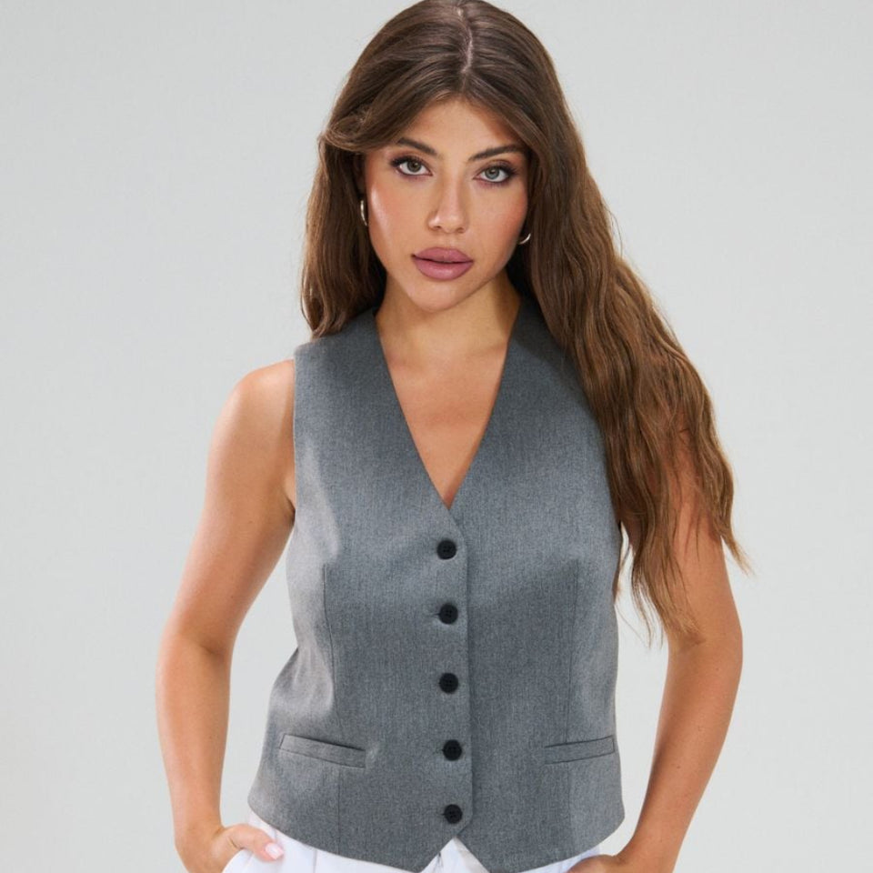 A woman modeling a sleeveless gray vest with a V-neck and black buttons, paired with white pants, against a light gray studio background.