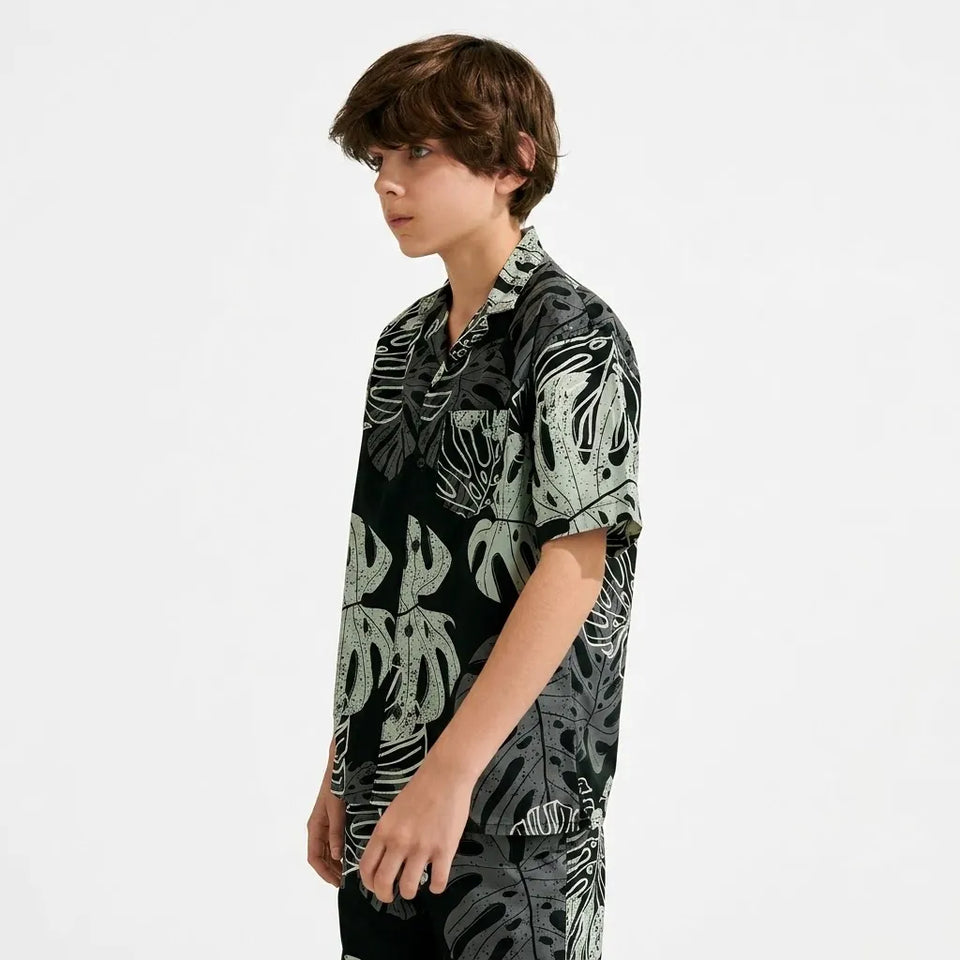 A young man with brown hair is shown in profile wearing a short-sleeve button-up shirt with a black base and a light gray tropical leaf pattern, set against a plain white studio background.