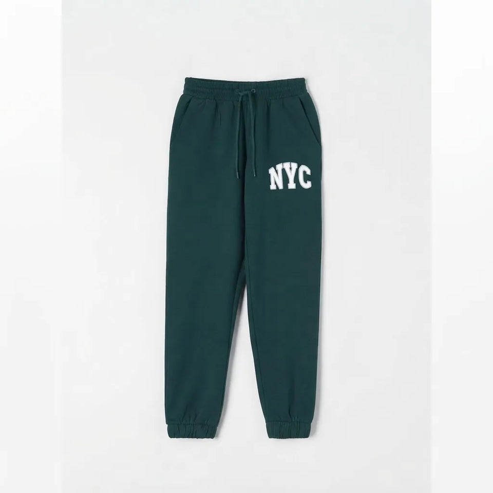 Dark green sweatpants with a white 'NYC' graphic on the left thigh, featuring an elastic waistband with a drawstring and elastic cuffs, shown against a light gray background.