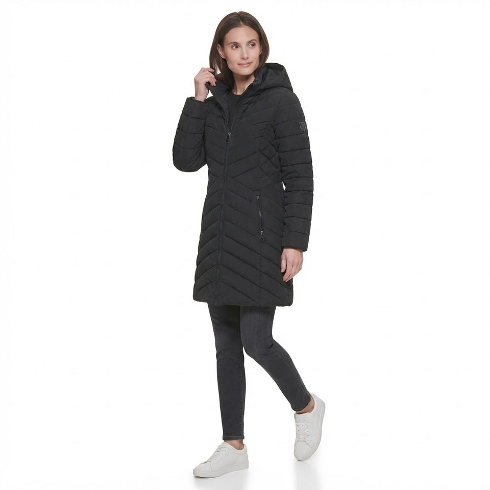 A woman modeling a black hooded quilted puffer coat with a chevron pattern, paired with dark gray leggings and white sneakers, against a solid white background.