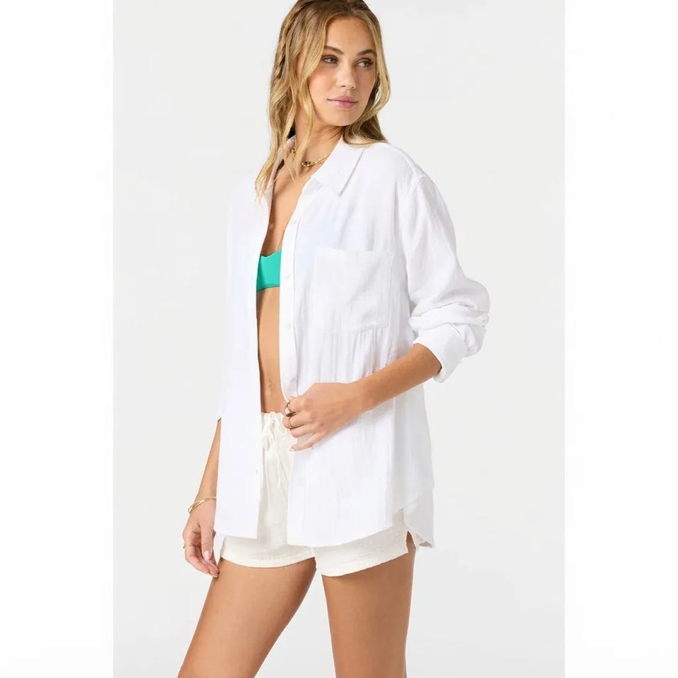 A woman modeling a white long-sleeve button-up shirt with a chest pocket, worn open over a turquoise bikini top and cream shorts, against a plain light gray studio background.