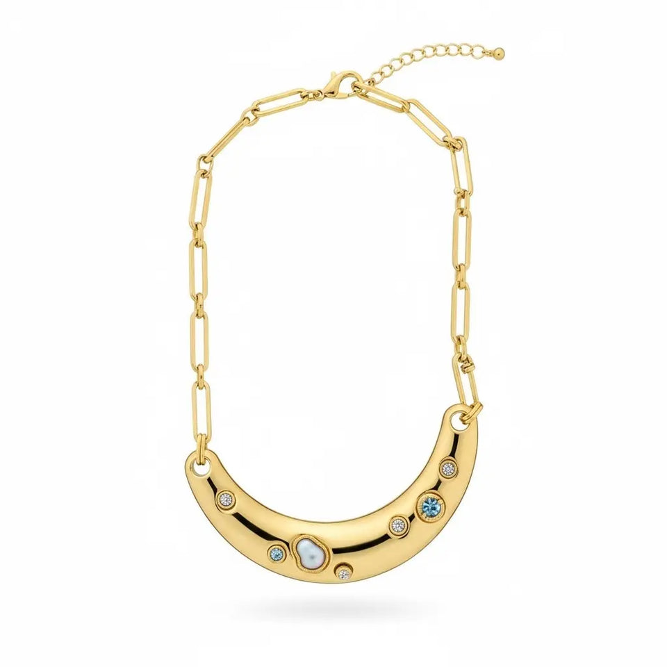 A gold crescent moon necklace with a paperclip chain, featuring small clear and blue gemstones set into the curve, shown on a plain white background.