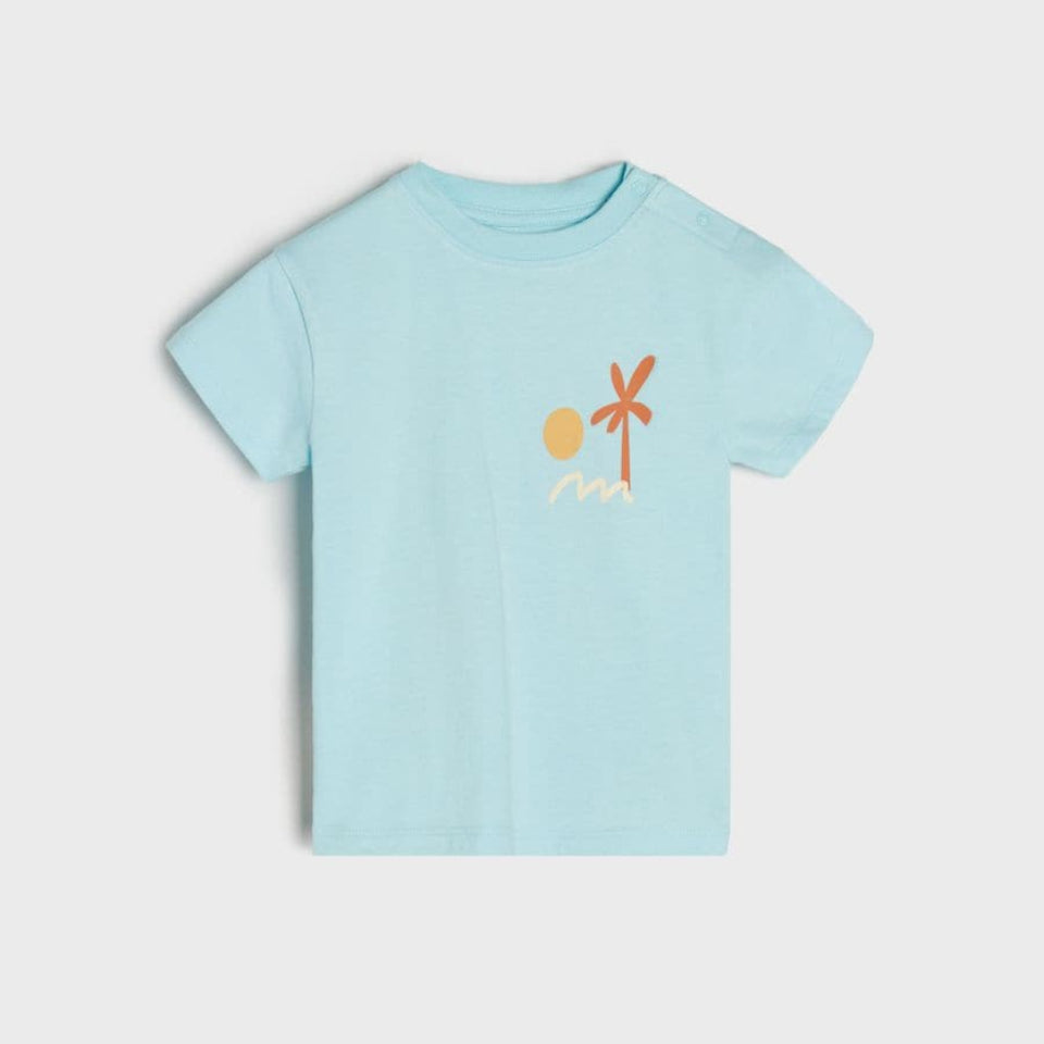 A light blue short-sleeve baby t-shirt with a small graphic of a yellow sun, orange palm tree, and white waves on the chest, displayed against a light gray background.