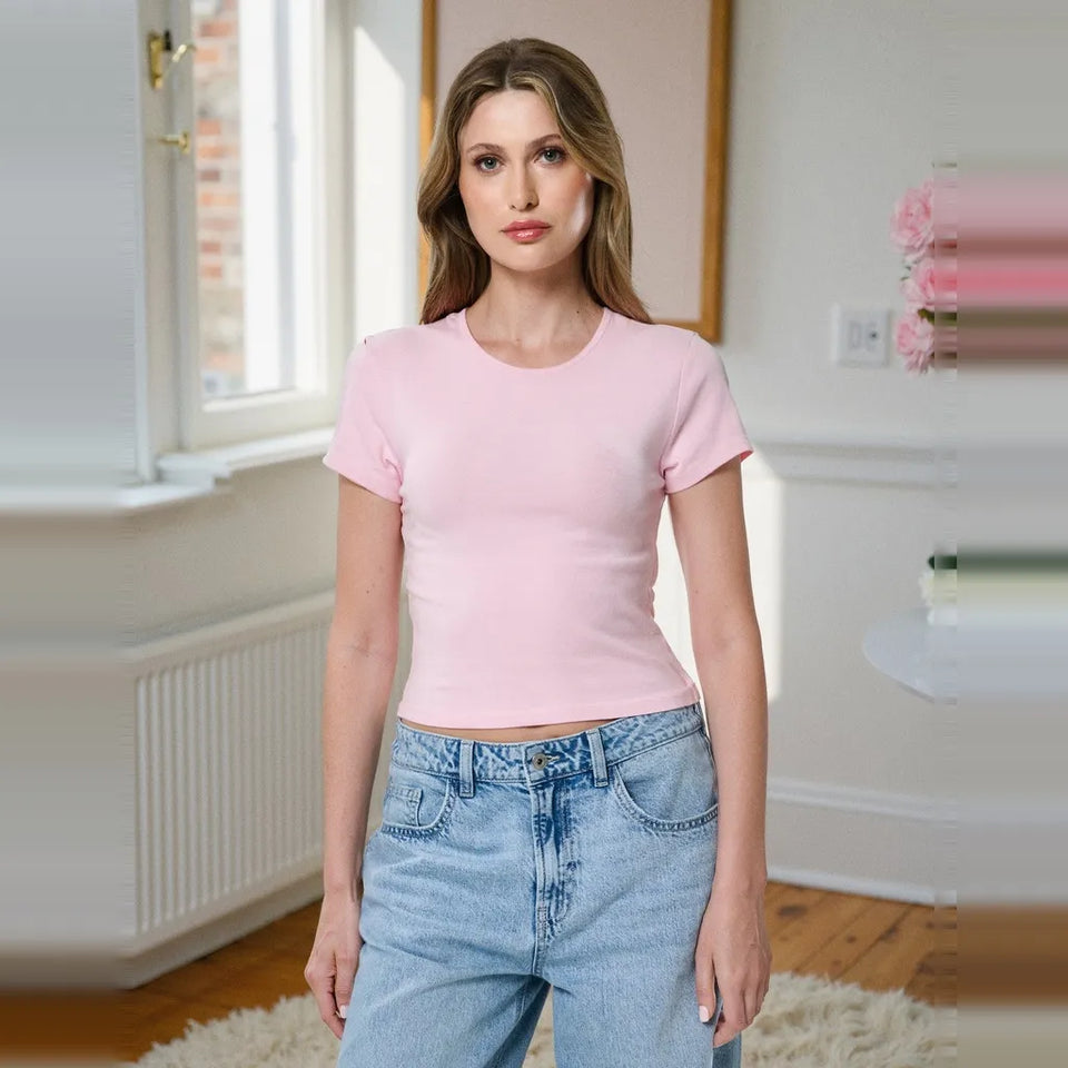 A woman modeling a fitted light pink short-sleeve crewneck t-shirt paired with light-wash blue jeans in a bright, modern room with white walls and a window.
