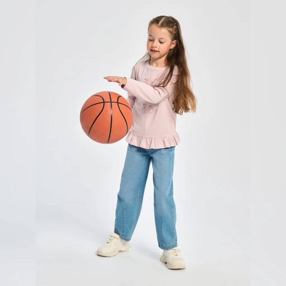 A young girl with long light brown hair stands on a light gray studio background, wearing a light pink long-sleeve ruffled shirt, blue jeans, and white sneakers while holding a basketball.