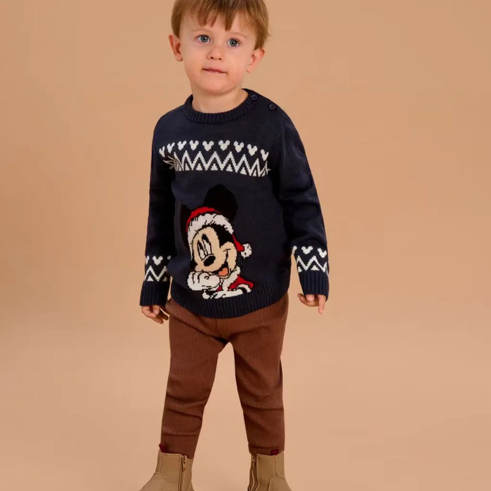 A toddler standing against a solid beige background, wearing a navy blue knit sweater with a white Nordic pattern and a Mickey Mouse graphic in a Santa hat, paired with brown pants and tan boots.