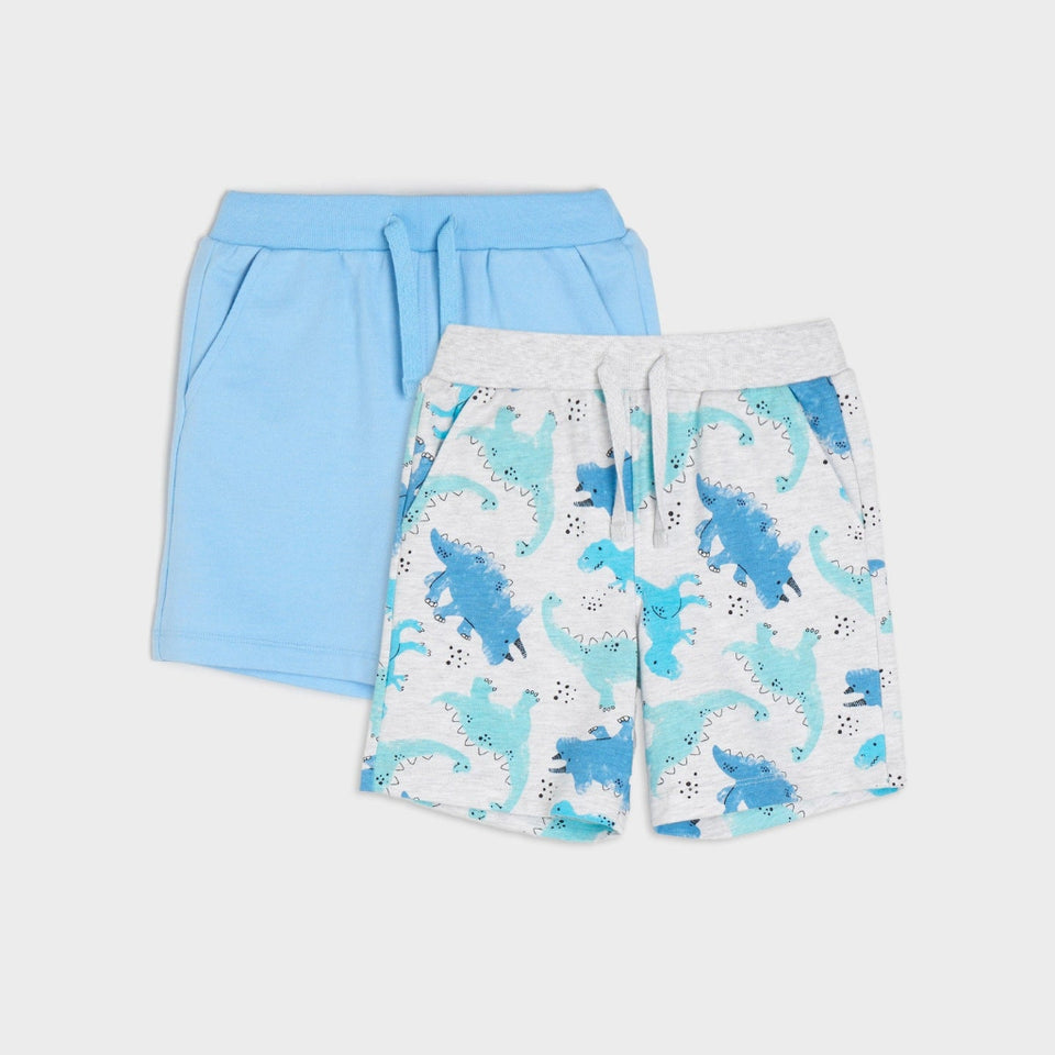 Two pairs of children's shorts laid flat on a light gray background. The back pair is solid light blue with a drawstring waist, and the front pair is white with a blue dinosaur pattern and a gray elastic waistband.