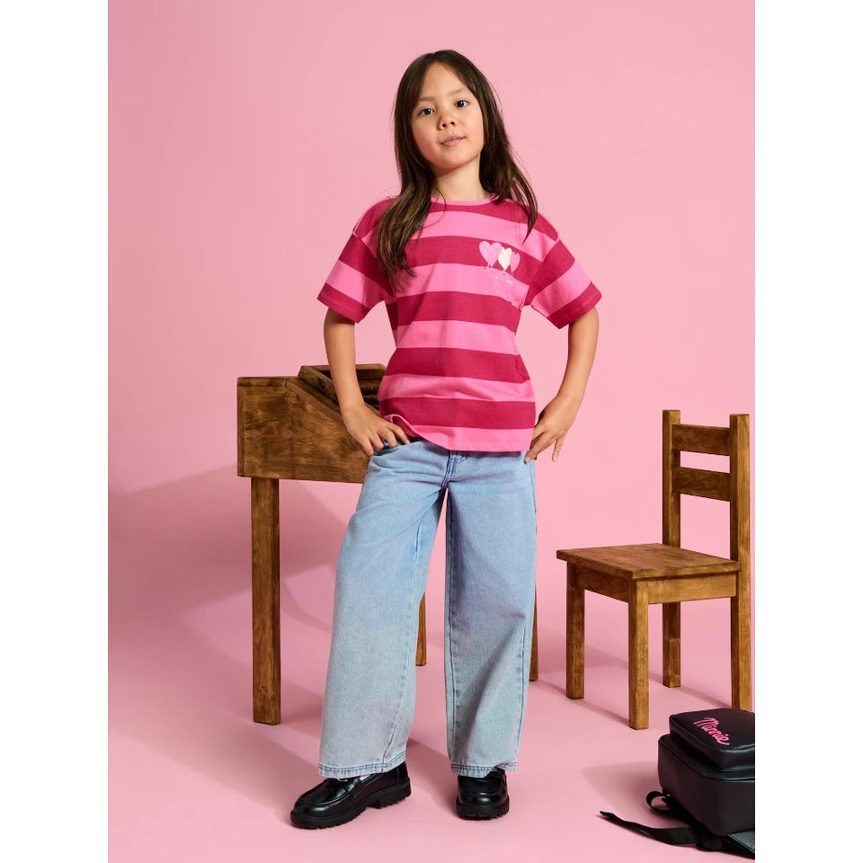 A young girl stands in a studio setting wearing a pink and magenta striped t-shirt with a small white heart graphic on the chest, paired with light-wash wide-leg jeans and black shoes. She is posed in front of a pink background with a wooden table and chair.