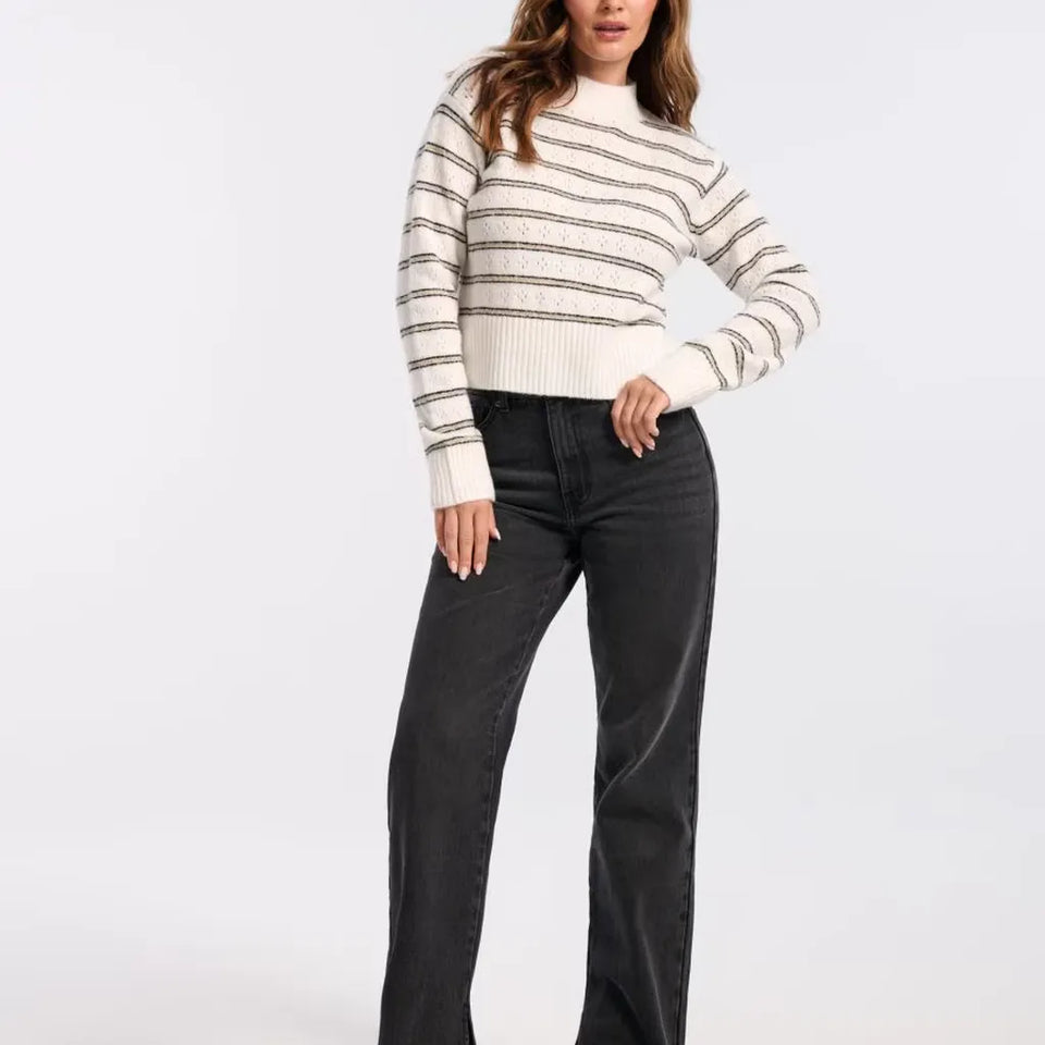 A woman modeling a cream knit sweater with thin dark horizontal stripes and black wide-leg jeans against a plain light gray studio background.