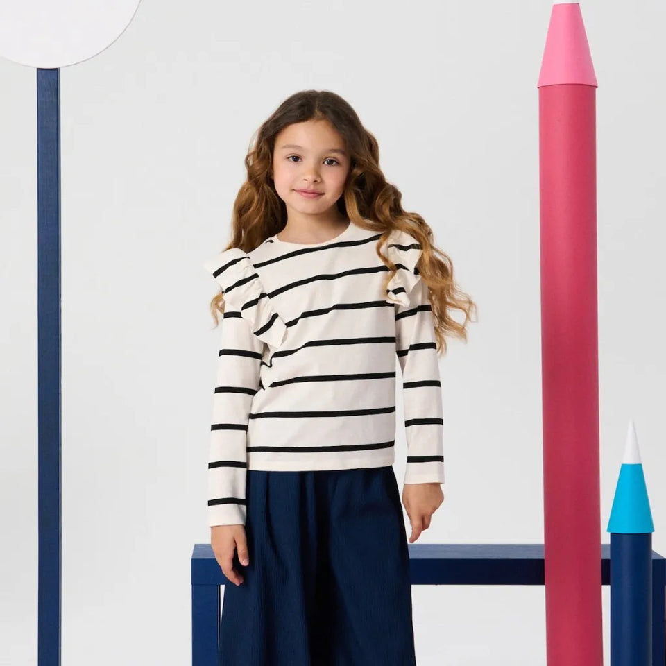 A young girl with long wavy brown hair stands in a studio setting wearing a cream long-sleeve top with black horizontal stripes and ruffle details on the shoulders, paired with dark navy wide-leg pants. She is framed by large, stylized pink and blue pencil props against a clean white background.