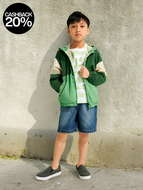 Child wearing a green jacket and denim shorts standing against a concrete wall with a cashback offer graphic.