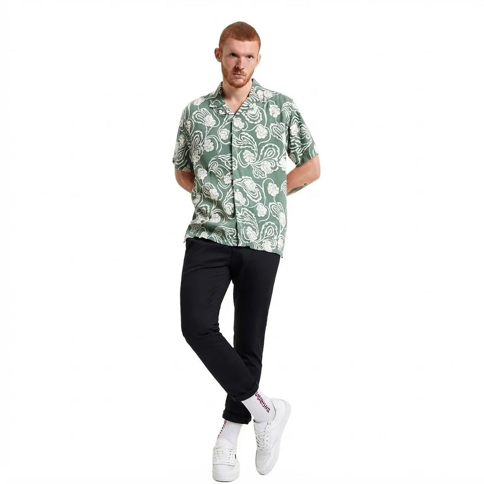 A man modeling a short-sleeve button-up shirt with a green and white paisley pattern, paired with black pants and white sneakers, against a plain white background.