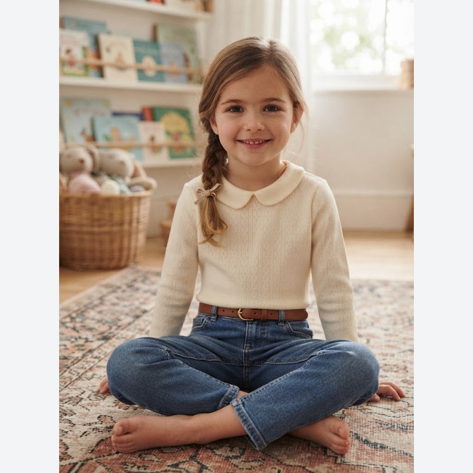 A young girl with a braided ponytail sits cross-legged on a patterned rug in a child's room, wearing a cream knit long-sleeve top with a Peter Pan collar, blue jeans, and a brown belt.