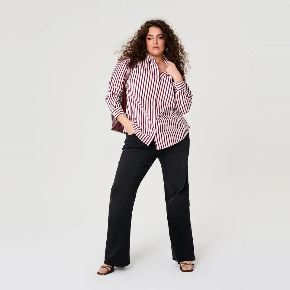 A woman modeling a red and white vertically striped button-down shirt with black wide-leg pants, standing against a plain light gray studio background.