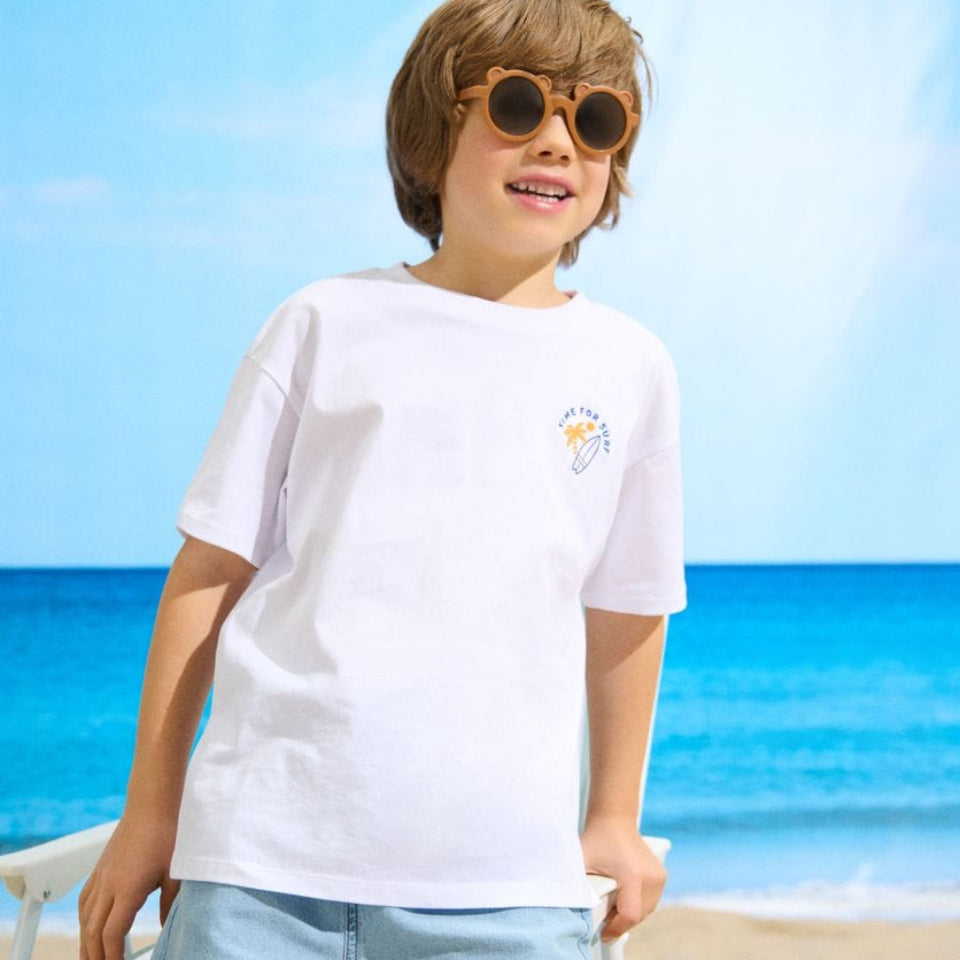 A child wearing a white short-sleeve t-shirt with a small yellow and blue graphic on the left chest, paired with light blue denim shorts, standing on a sandy beach with the ocean and blue sky in the background.
