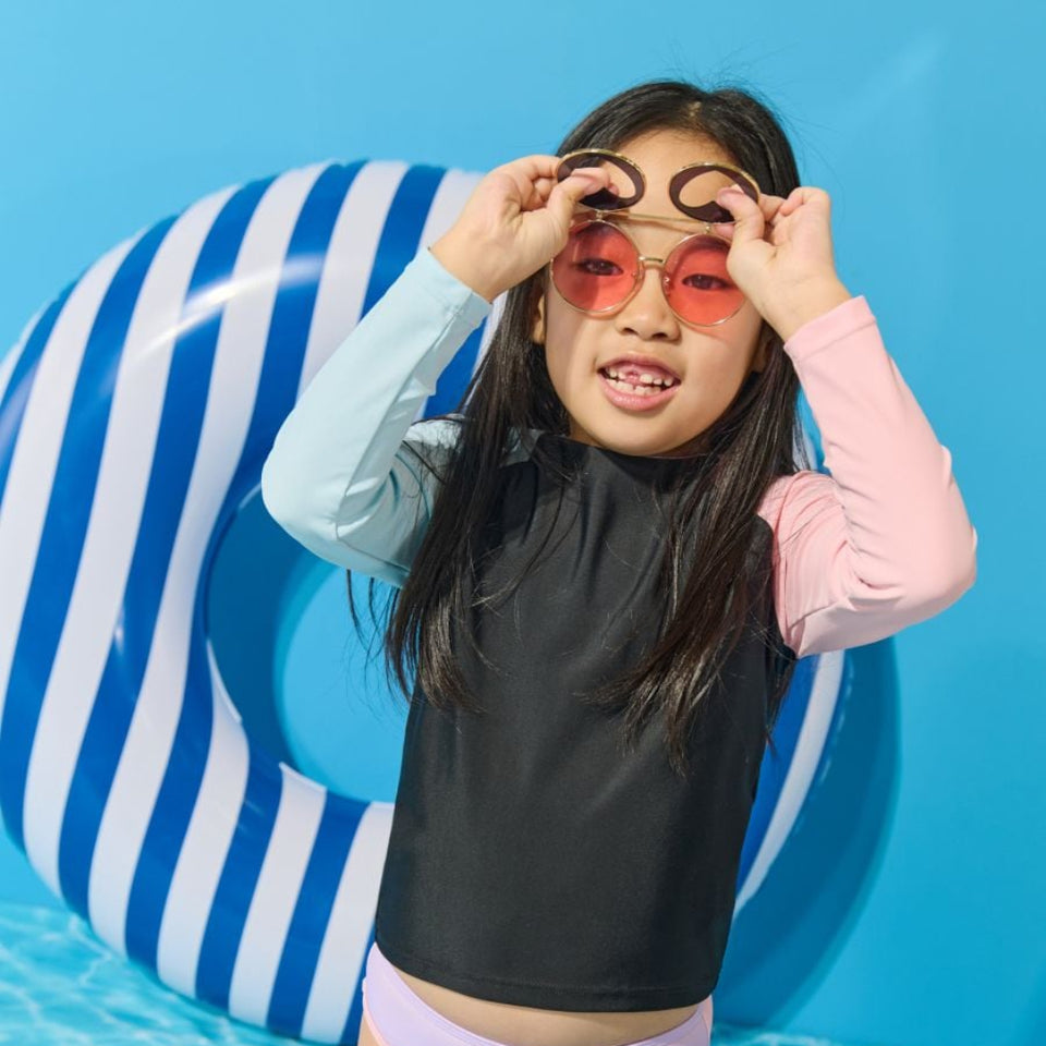 A young girl wearing a two-tone long-sleeve swim shirt with a black front panel and pastel blue and pink sleeves, paired with lavender swim bottoms, posing in a pool with a blue and white striped float.