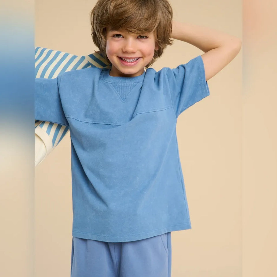 A smiling young boy wearing a light blue short-sleeve crewneck t-shirt and matching blue pants, posing against a warm beige background.