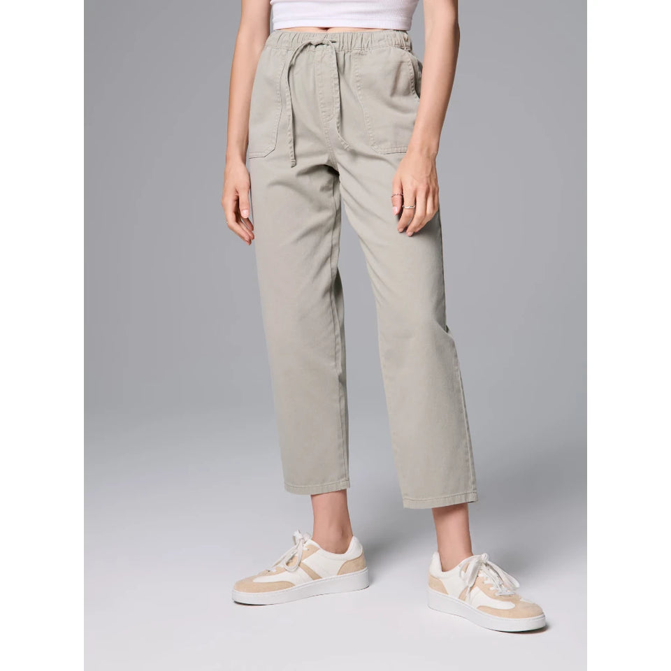 A model from the waist down wearing light beige cropped pants with a drawstring waist and side pockets, paired with beige and white sneakers, against a solid gray studio background.