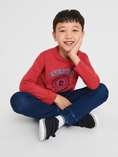 Child wearing a red sweatshirt with a logo and text, sitting on the floor against a white background