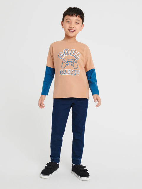 Child wearing a beige long-sleeve shirt with blue sleeves and navy pants on a white background