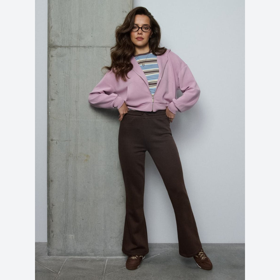A woman modeling a light pink zip-up hoodie over a striped t-shirt and dark brown wide-leg trousers, standing against a gray concrete wall.