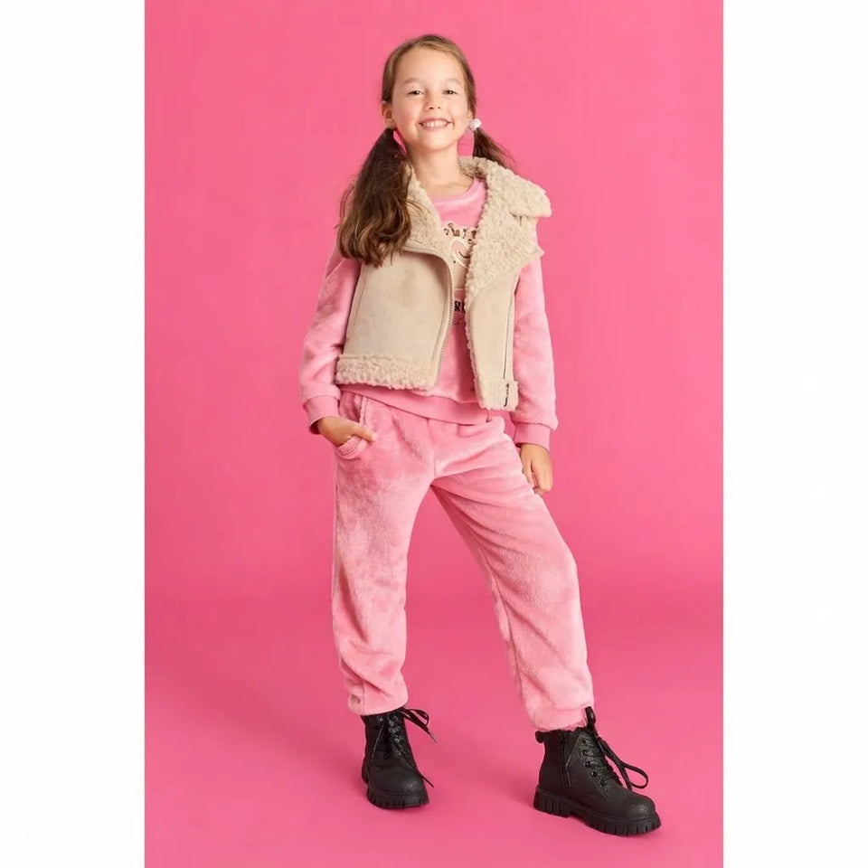 A young girl modeling a pink velour lounge set consisting of a long-sleeve top and matching sweatpants, styled with a beige sherpa-lined vest and black boots against a solid pink background.