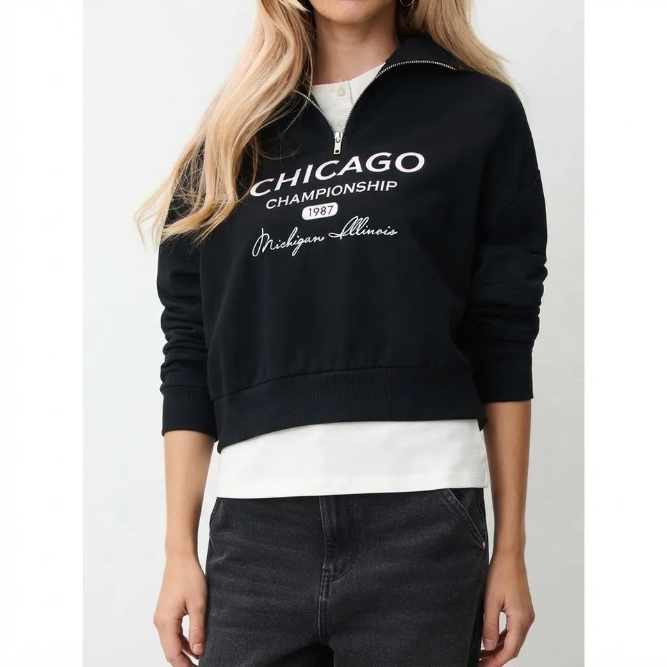 A woman wearing a black half-zip sweatshirt with 'CHICAGO CHAMPIONSHIP 1987 Michigan State' printed in white on the front, layered over a white t-shirt and dark gray jeans.
