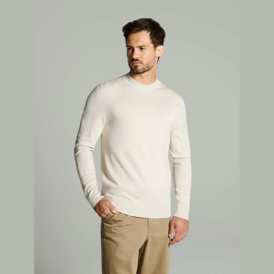 A man wearing a cream-colored long-sleeve mock neck sweater and khaki pants, standing against a solid light gray background.