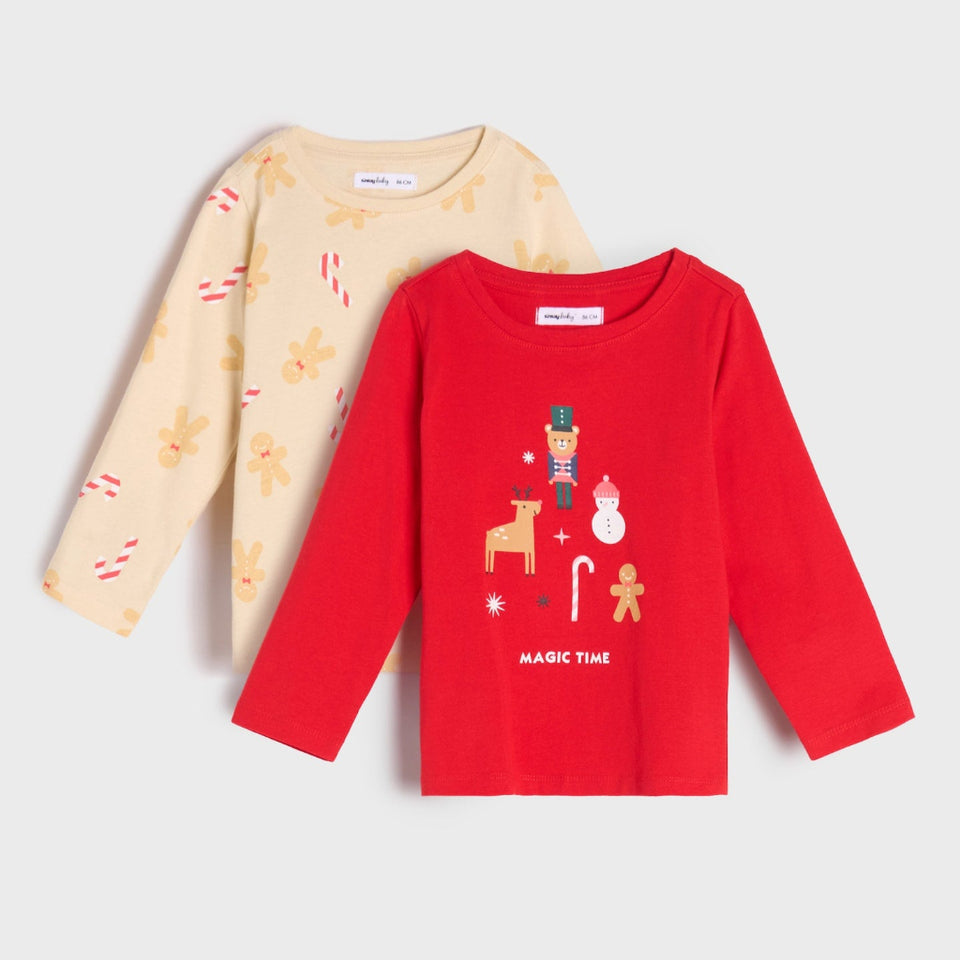 Two long-sleeve children's shirts displayed on a light gray background. The front shirt is bright red with a Christmas-themed graphic print featuring a nutcracker, reindeer, candy cane, snowman, gingerbread man, and the text 'MAGIC TIME'.