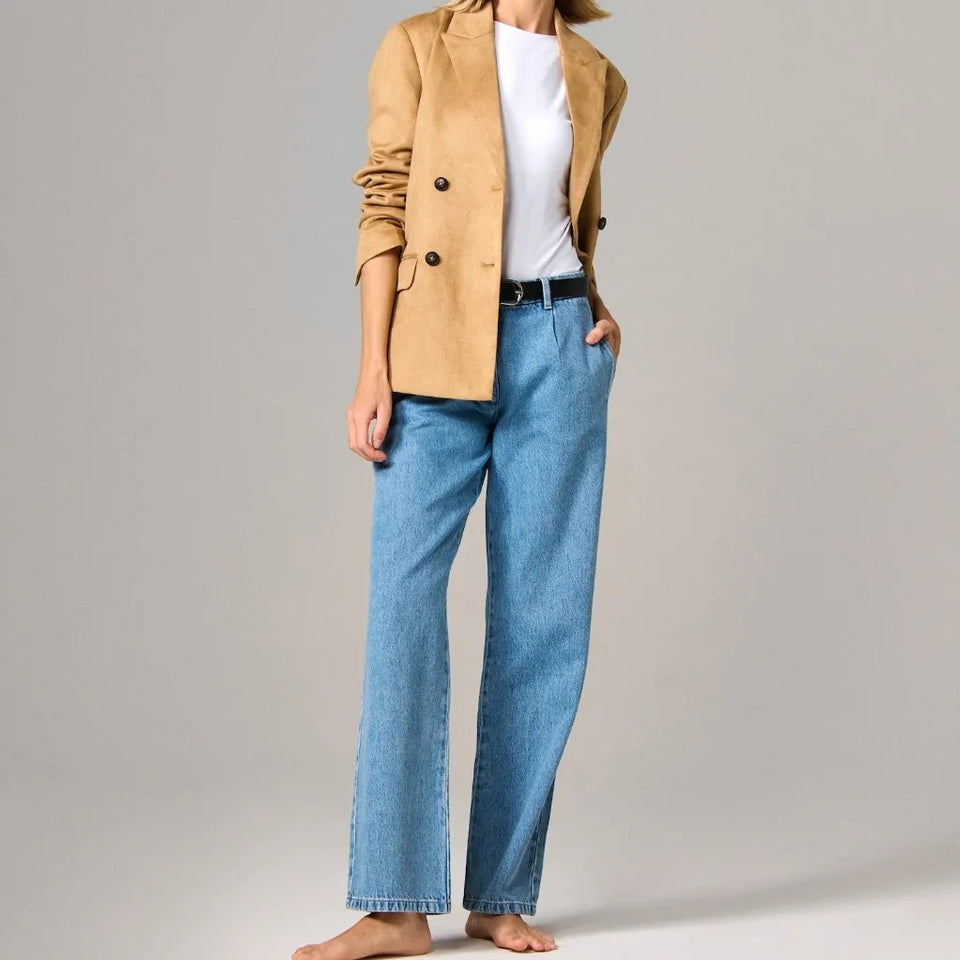 A model wearing light-wash blue straight-leg jeans, a white t-shirt, and a tan suede blazer, standing barefoot against a neutral gray studio background.