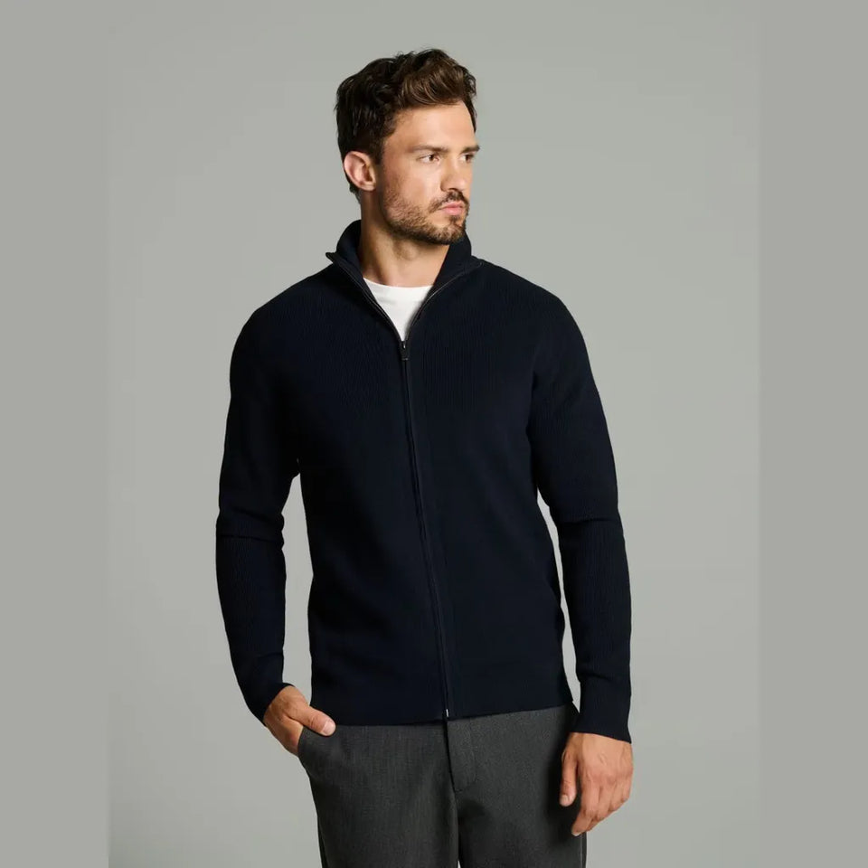A man wearing a dark navy blue zip-up sweater over a white t-shirt and gray trousers, standing against a solid gray studio background.