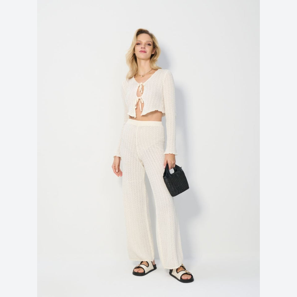 A woman modeling a cream-colored ribbed knit two-piece set consisting of a long-sleeve crop top with front tie details and matching wide-leg pants, paired with black sandals and a small black handbag against a light gray studio background.