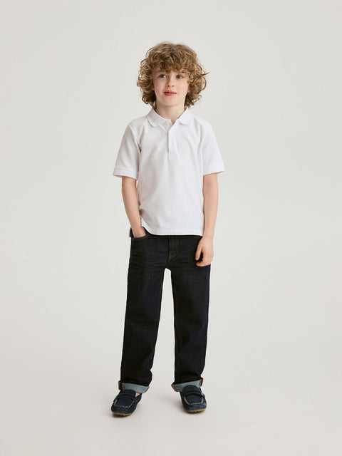 Young boy wearing a white polo shirt and black jeans on a light gray background