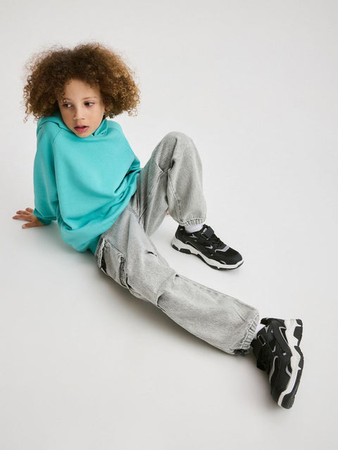 Child sitting on a white floor wearing a turquoise hoodie, gray pants, and black sneakers.
