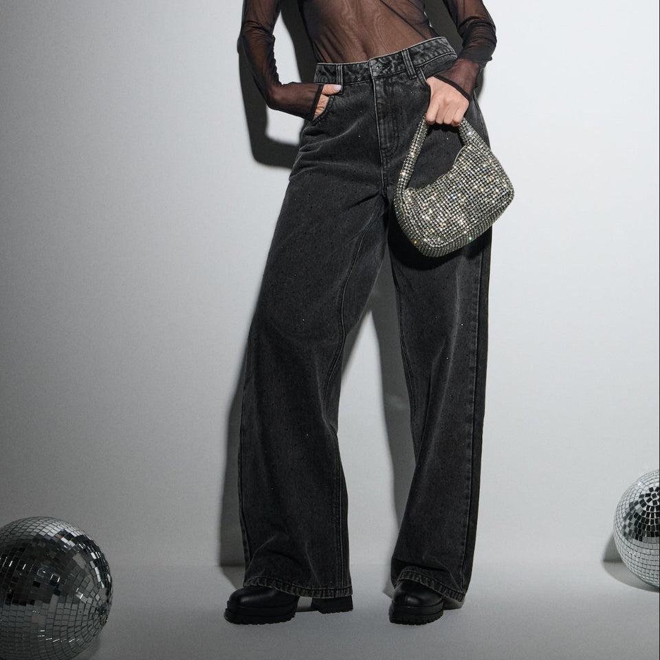A model stands against a light gray studio background wearing a sheer black long-sleeve crop top over a black bandeau, high-waisted wide-leg dark gray jeans, and holding a silver sequined handbag.