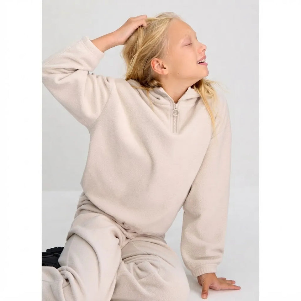 A young girl with blonde hair wearing a beige fleece half-zip hoodie and matching sweatpants, posing against a light gray background.