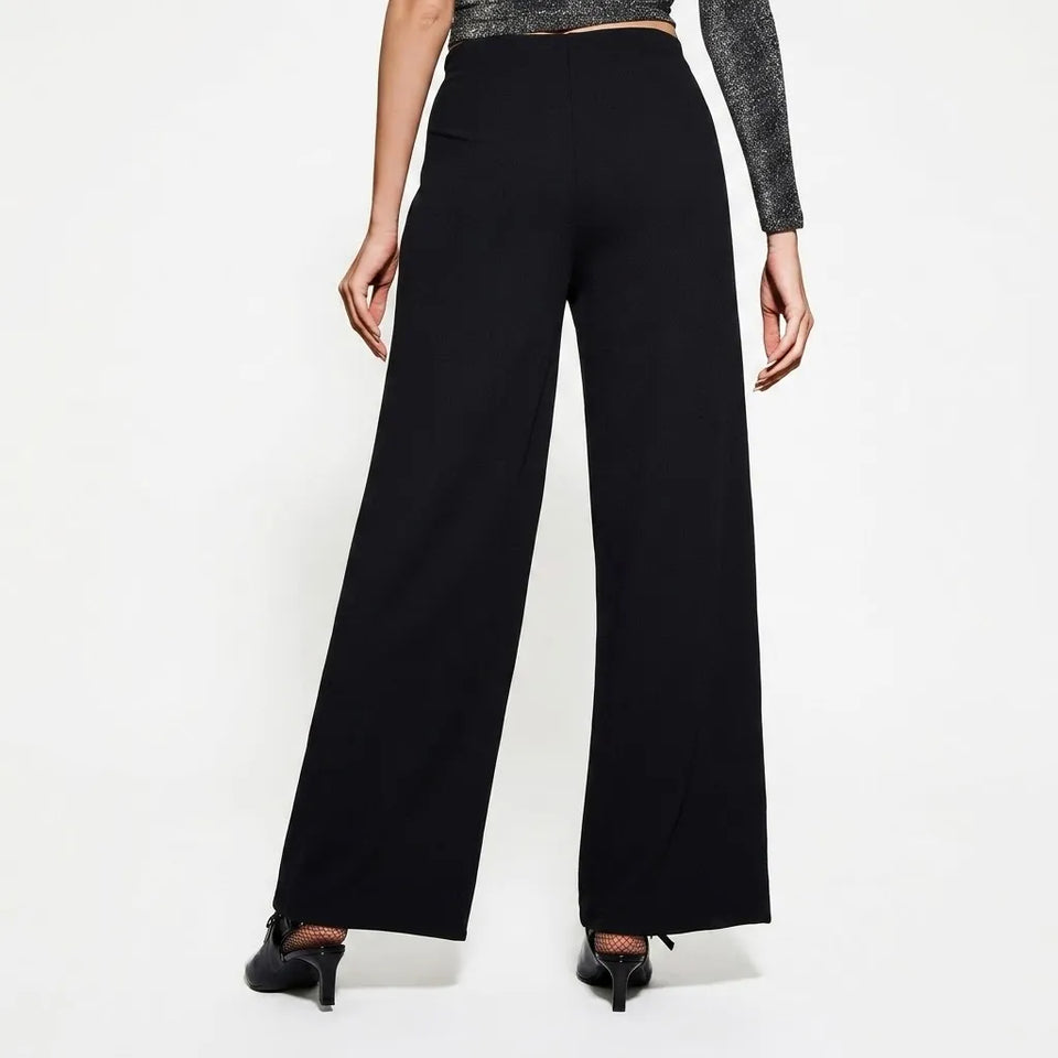 A model from the back wearing high-waisted, wide-leg black pants, a textured gray long-sleeve crop top, and black pointed-toe heels against a plain white studio background.