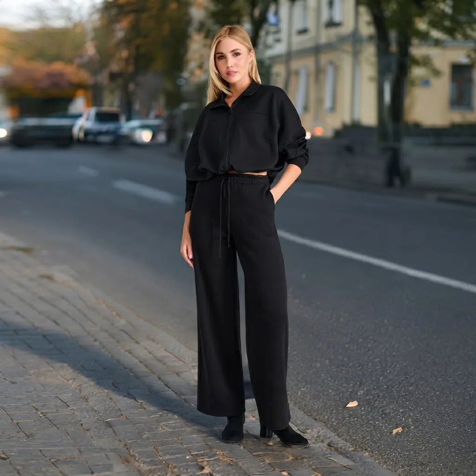 A woman stands on a city street wearing a black long-sleeve collared top and matching wide-leg trousers with a drawstring waist, paired with black ankle boots.