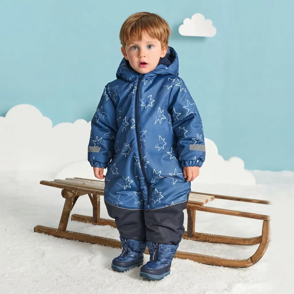 A young child wearing a blue hooded snowsuit with a white star pattern, dark pants, and blue boots, standing in front of a wooden sled against a light blue backdrop with white cloud decorations.
