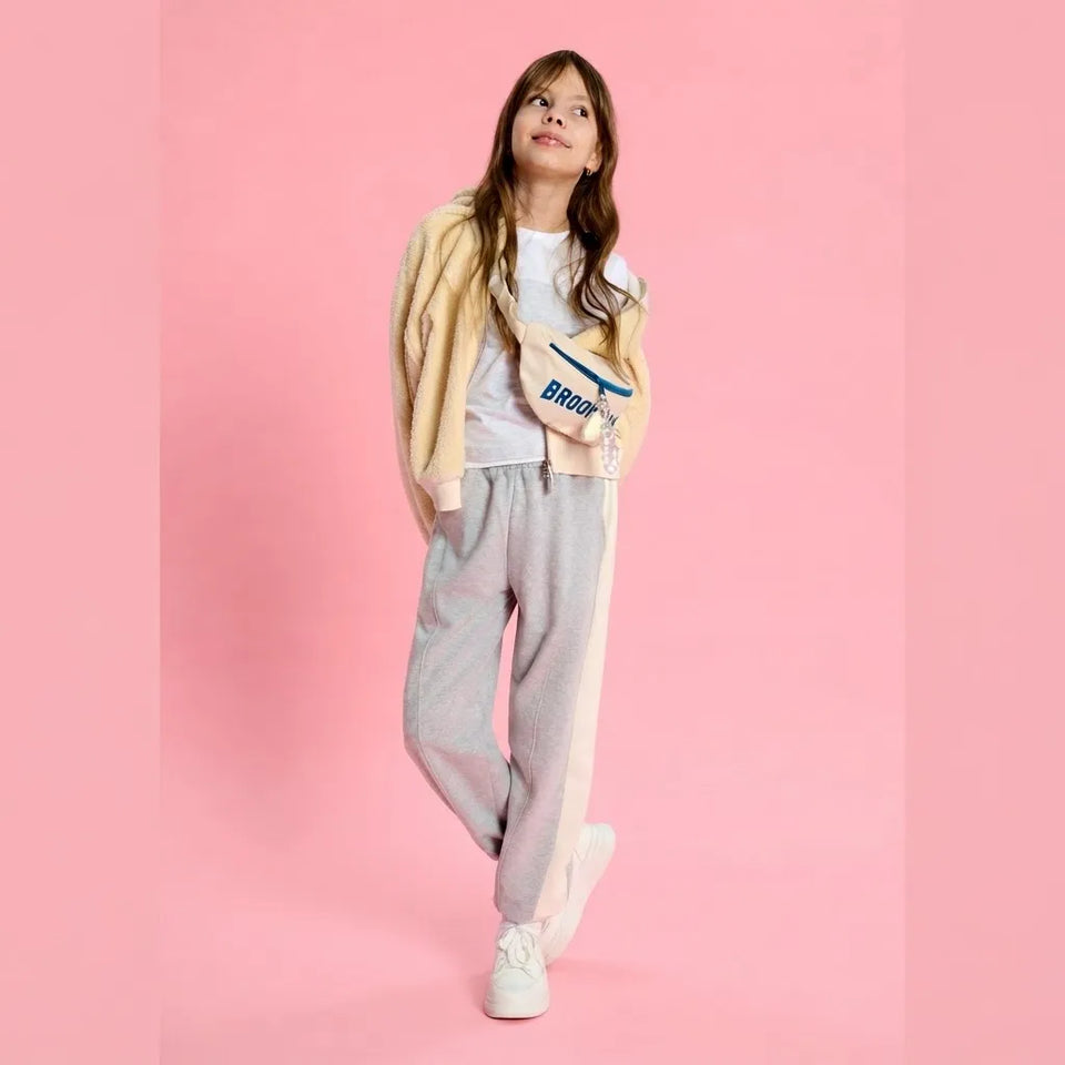 A young girl models a casual streetwear outfit consisting of a beige cardigan, white t-shirt, light gray sweatpants with a pink stripe, and white sneakers, set against a solid pink background.