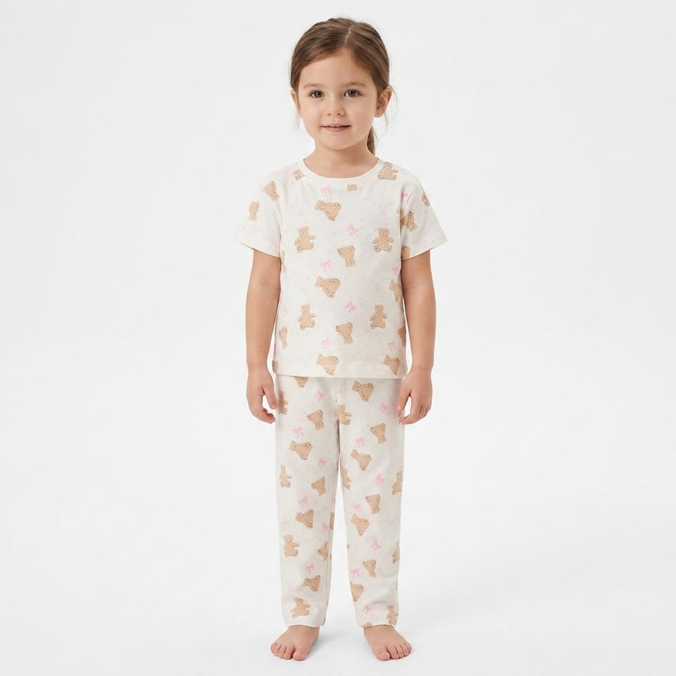 A young girl stands against a plain white studio background wearing a matching short-sleeve pajama top and pants set in cream with a repeating teddy bear print in tan and small pink bows.