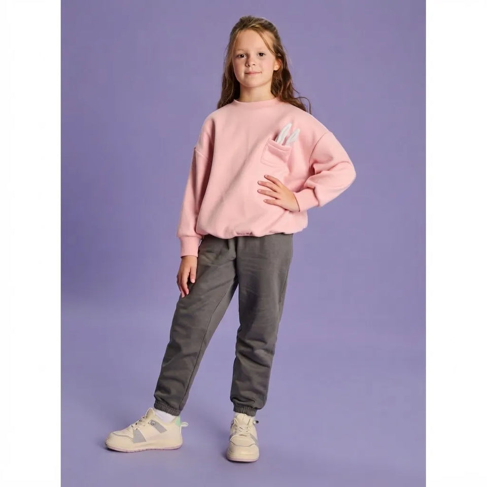 A young girl modeling a light pink crewneck sweatshirt with a front pocket holding white pens, paired with dark gray jogger pants and white sneakers, set against a solid lavender studio background.