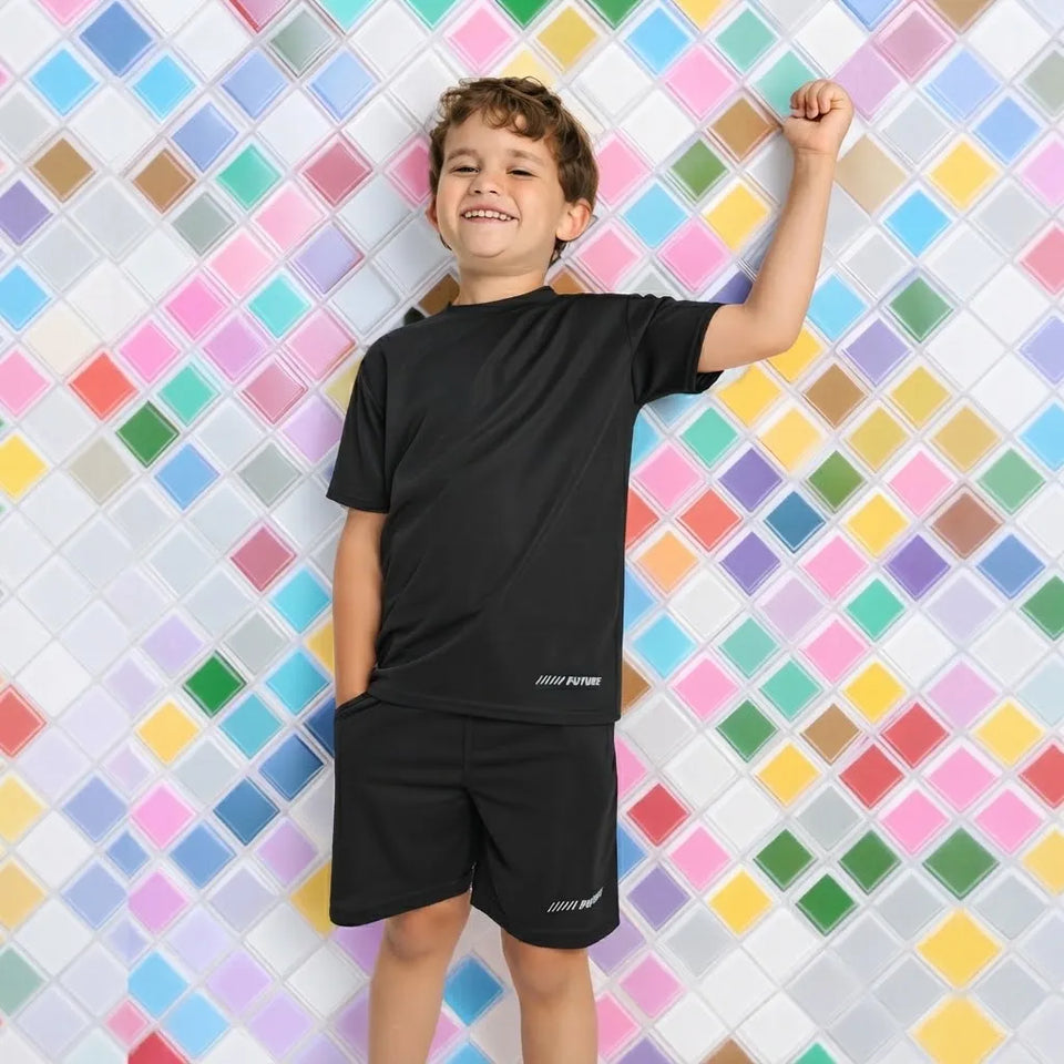 A young boy smiling and posing in a black athletic t-shirt and matching shorts set, standing against a colorful diamond-patterned tiled wall.