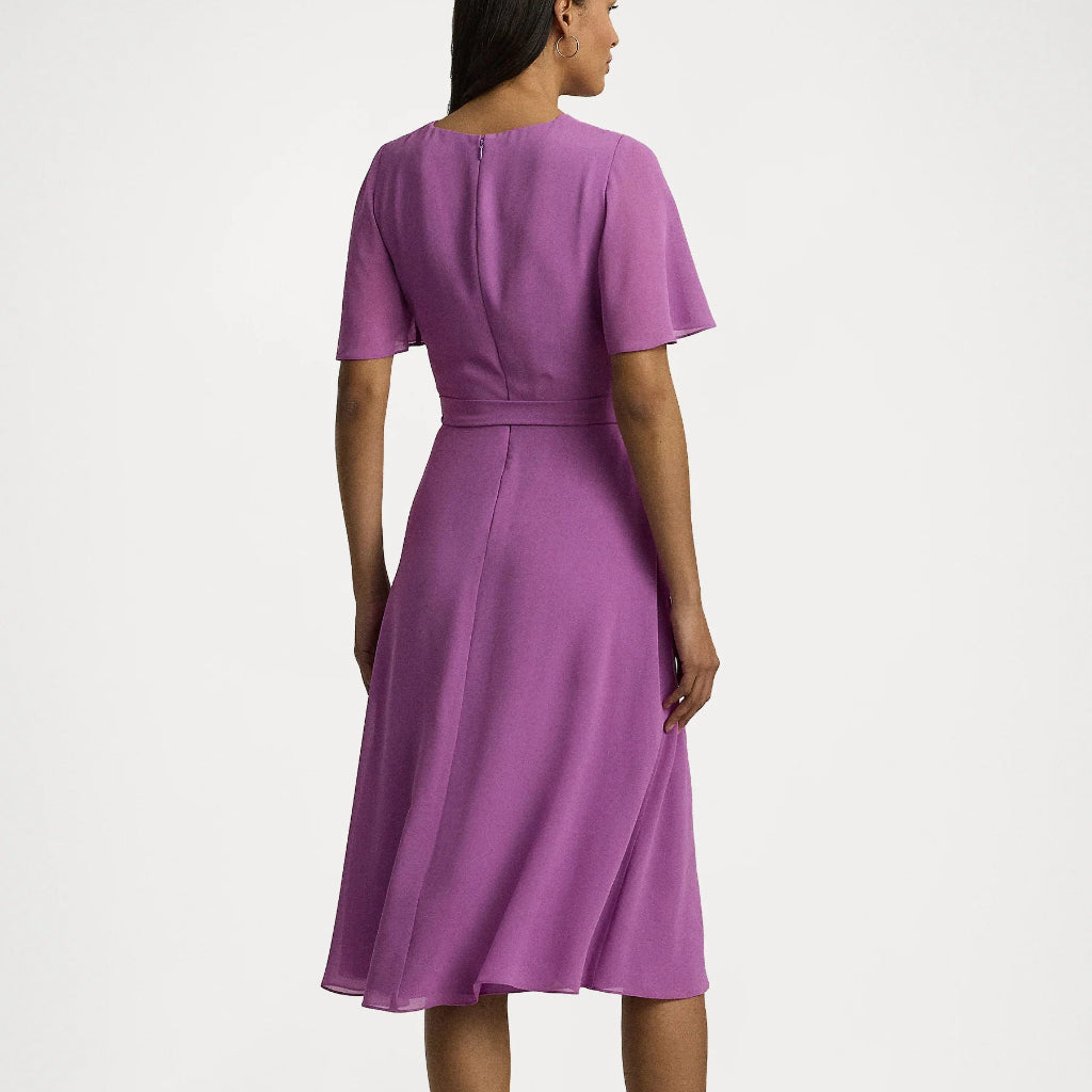 A woman shown from the back wearing a short-sleeved purple midi dress with a defined waist and black strappy heels, photographed against a plain light gray studio background.