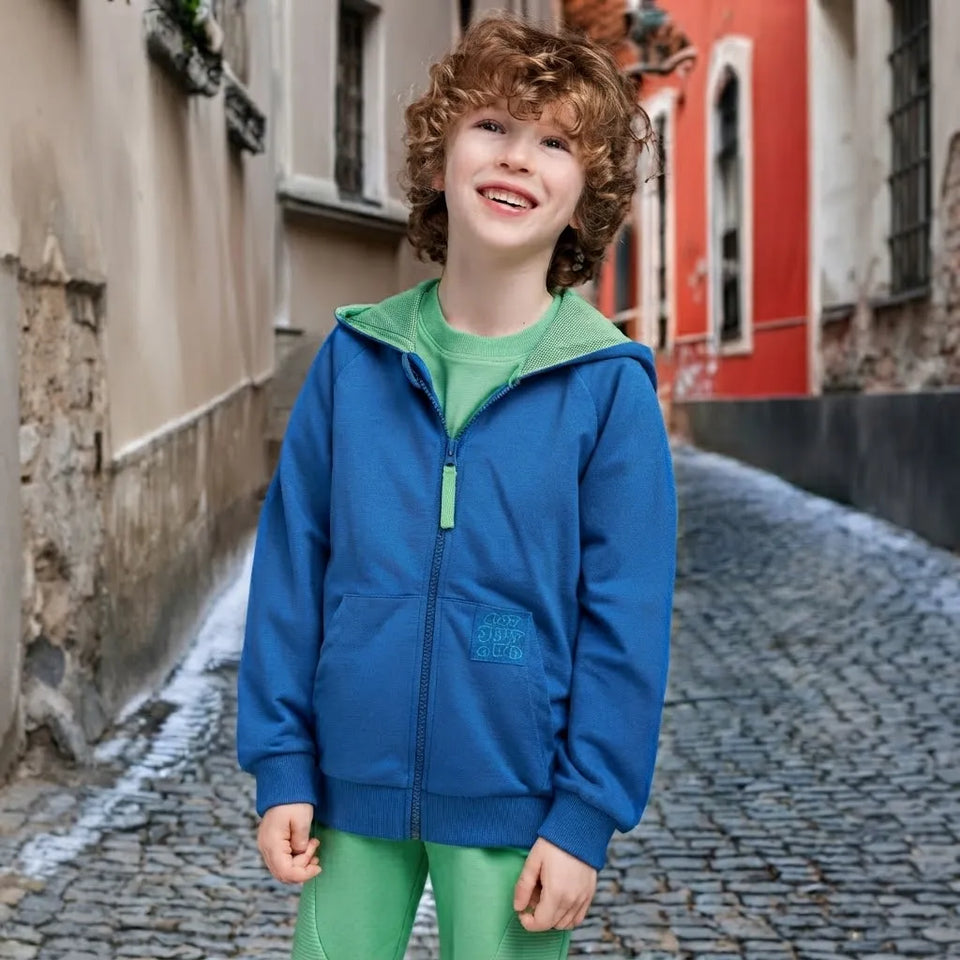 A smiling young boy with curly light brown hair stands on a cobblestone street wearing a blue zip-up hoodie with a green inner hood lining and green leggings.