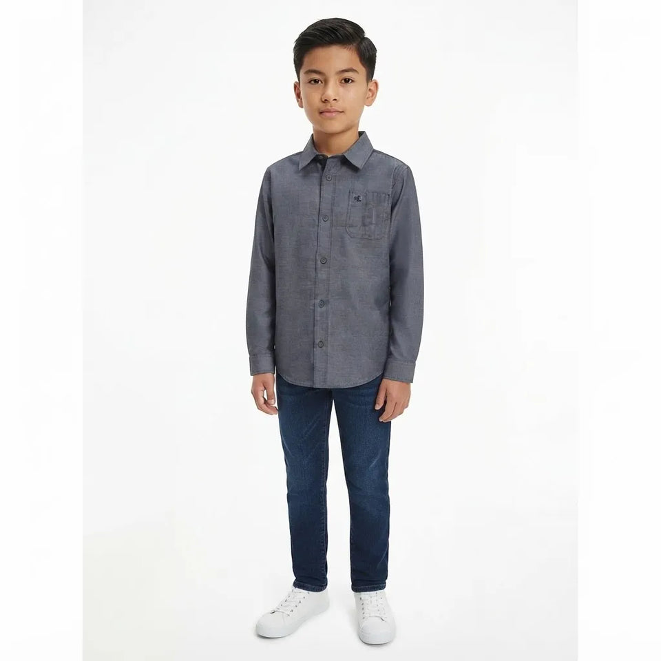 A young boy modeling a long-sleeve gray button-up shirt with dark blue jeans and white sneakers, standing against a plain white studio background.