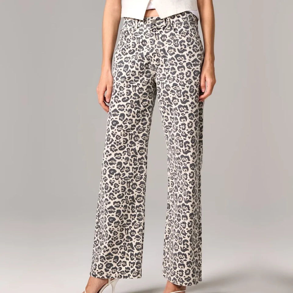 A model wearing high-waisted, wide-leg trousers with a gray and black leopard print on a light beige background, paired with a white top and clear high heels, set against a neutral gray studio background.
