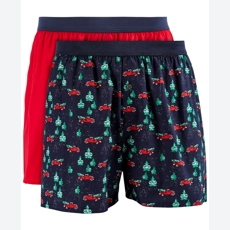 Two pairs of men's boxer briefs shown overlapping on a white background. The front pair has a dark navy base with a repeating holiday print of red pickup trucks and green Christmas trees, and the back pair is solid bright red with a navy waistband.