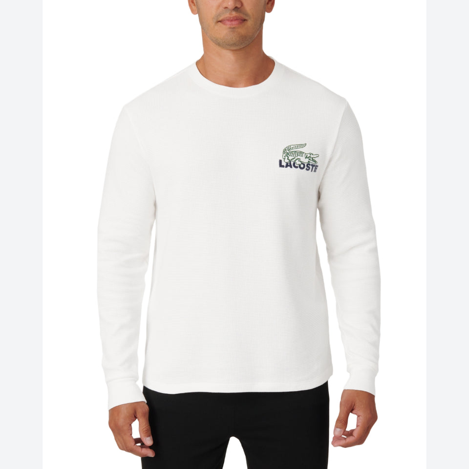 A man wearing a white long-sleeve crewneck sweatshirt with a green and navy Lacoste crocodile logo on the left chest, paired with black pants, against a plain white background.