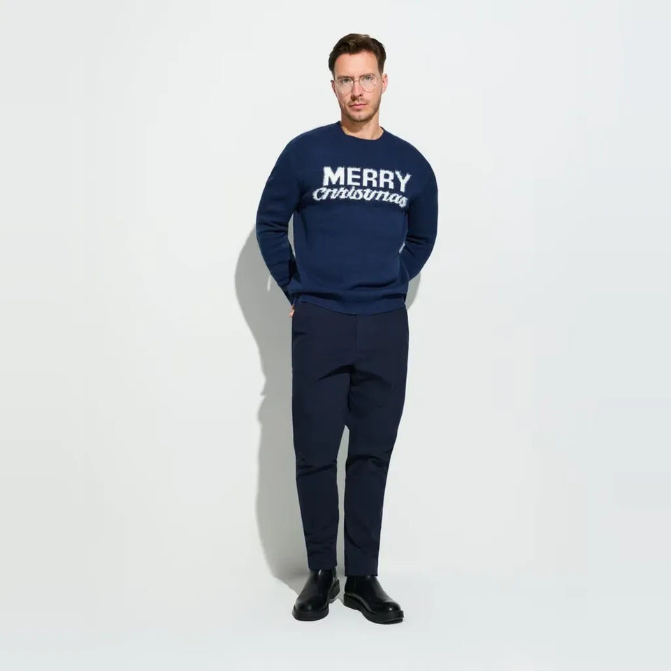 A man wearing a navy blue crewneck sweatshirt with 'MERRY christmas' printed in white across the chest, paired with navy trousers and black shoes, standing against a plain white studio background.